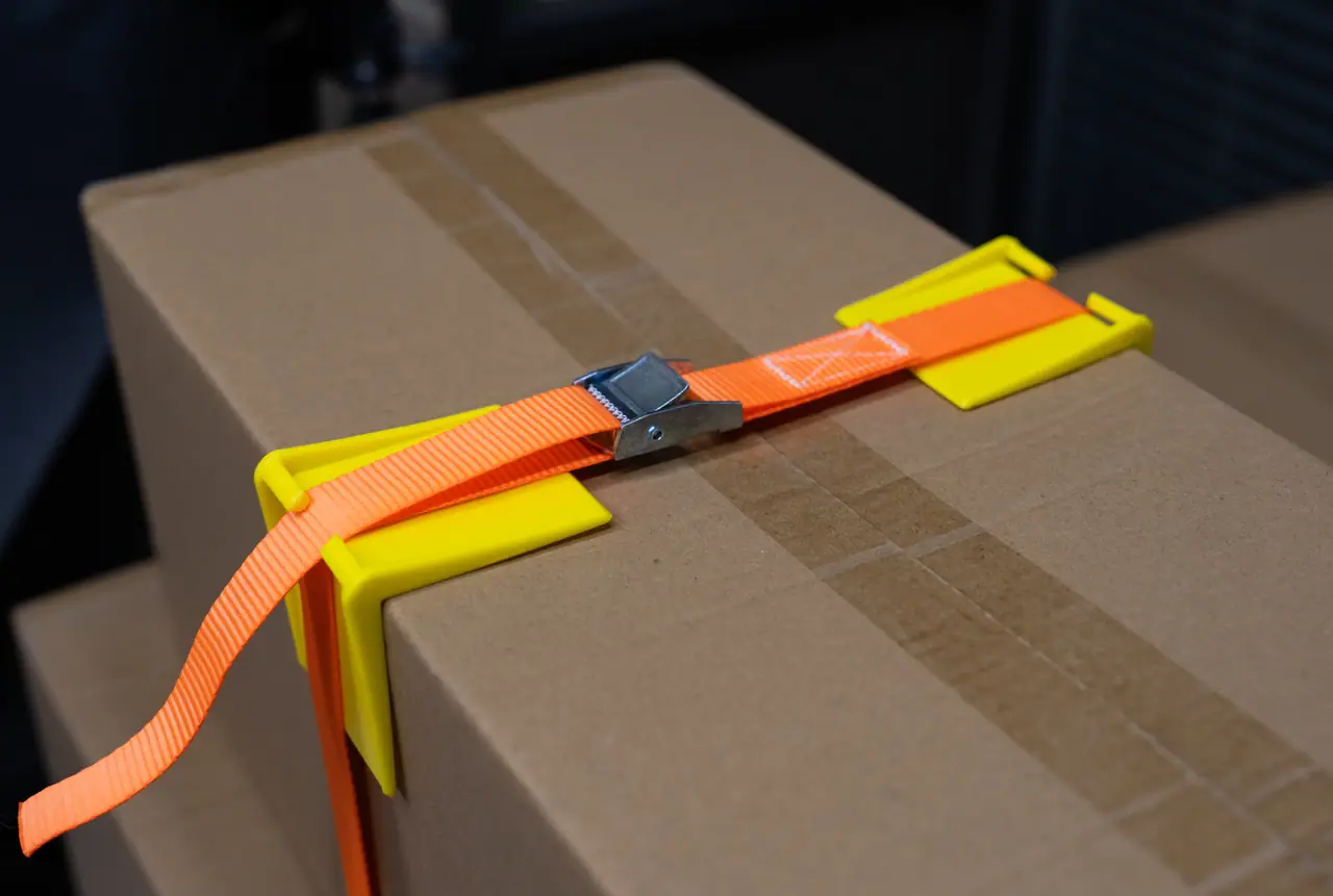 Sangle à boucle à came passée dans des protections d’angle en ABS jaunes sur un carton